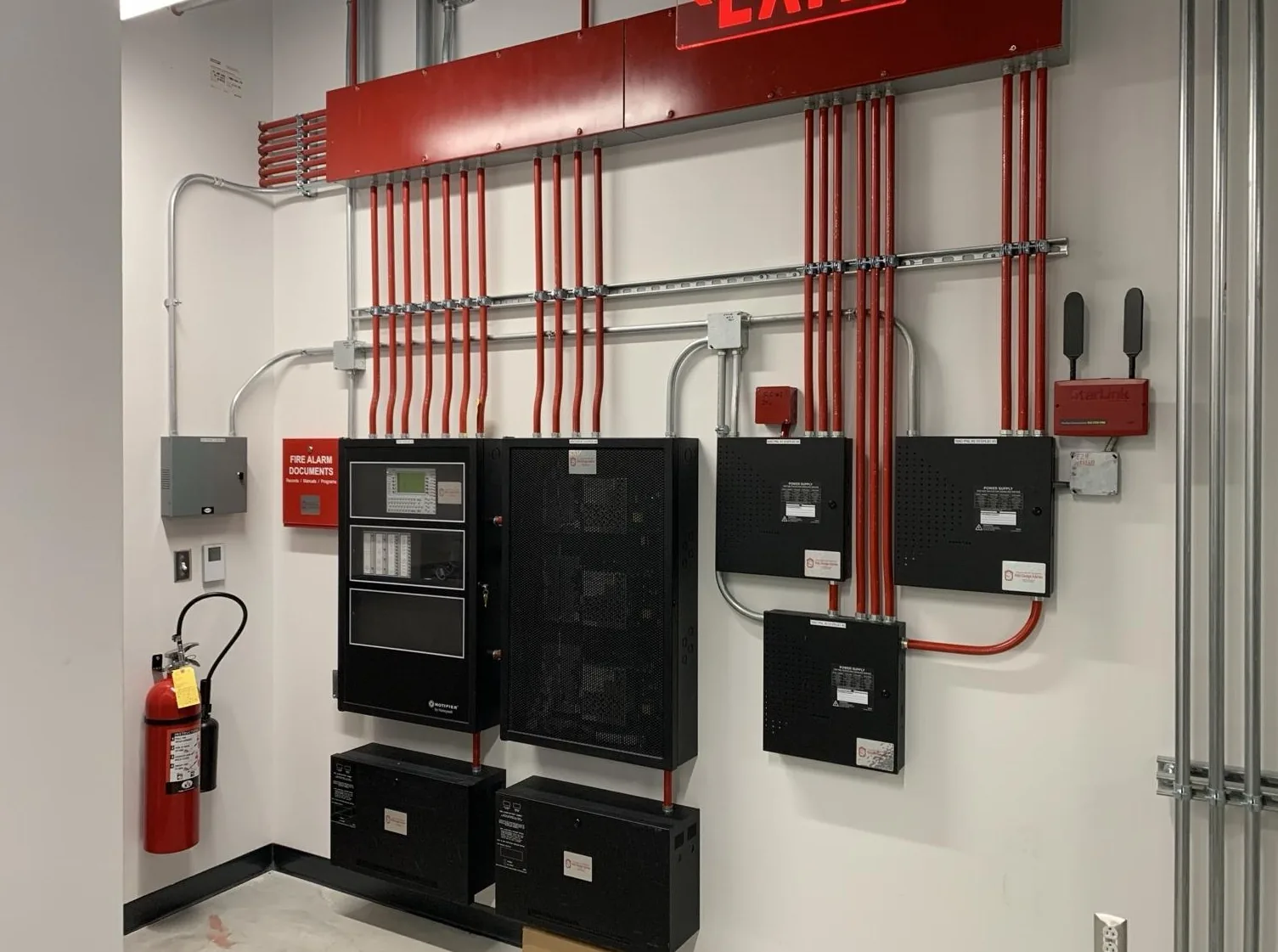 A commercial equipment wall with red conduit runs dropping into cabinets beneath an exit sign